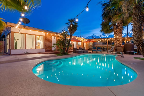 Illuminated backyard pool with brick house, palm trees, string lights, and lounge seating at dusk