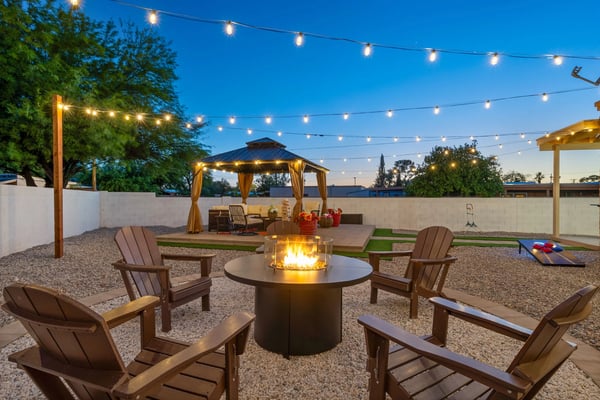Backyard patio at dusk with fire pit, Adirondack chairs, string lights, gazebo, and cornhole game